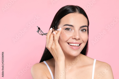 Beautiful young woman with eyelash curler on light pink background