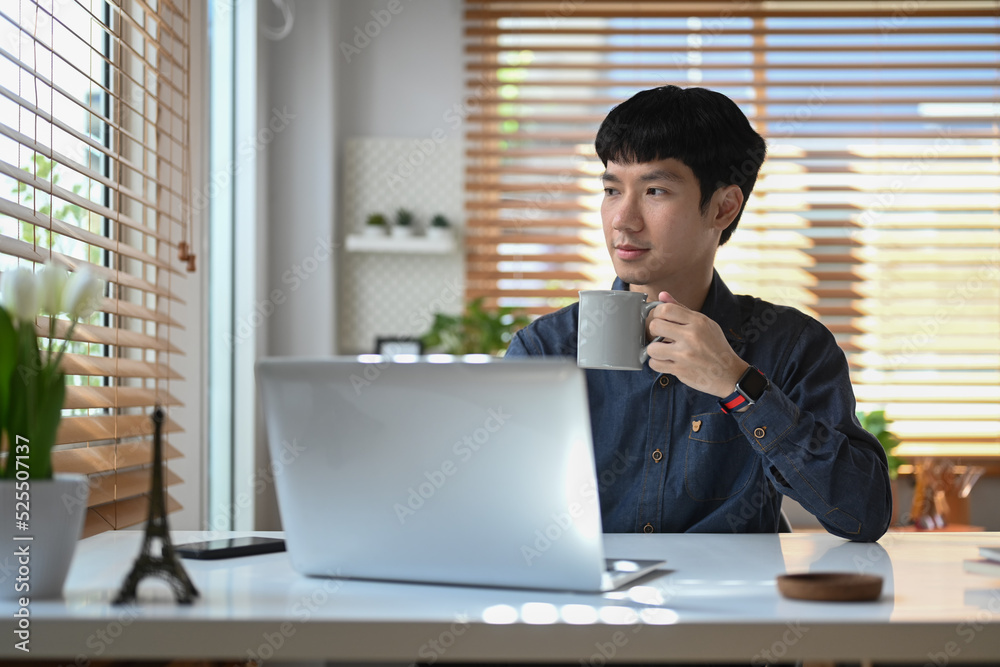 Thoughtful young businessman drinking coffee coffee and looking through window , spending free time in morning at his home office Thoughtful young businessman drinking coffee coffee and looking through window , spending free time in morning at his home office