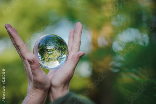 Fototapeta Naklejka Na Ścianę i Meble -  hands holding transparent glass ball or  crystal who reflects trees of a forest outside in green nature sphere symbolizing save the earth or planet