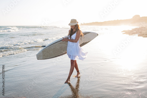 Young woman wearing a white dress walking with a surfboard through the sand