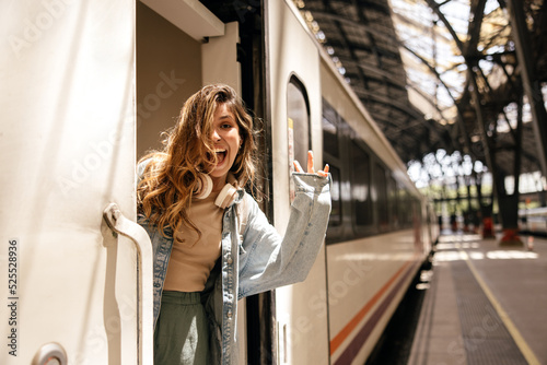 Joyful young caucasian woman sits in train carriage, waves her hand at camera. Blonde with wavy hair wears casual clothes. Mood, lifestyle, concept.