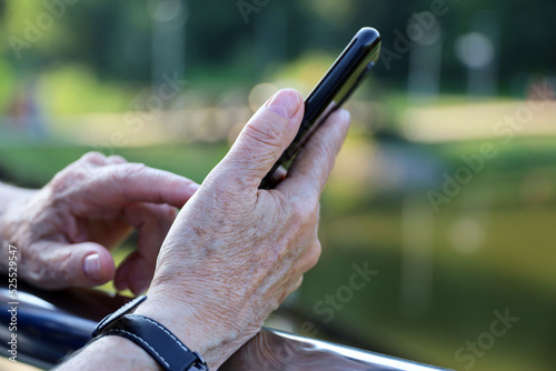 Wrinkled hands of elderly woman standing with smartphone in park. Mobile phone closeup, concept of online communication in retirement