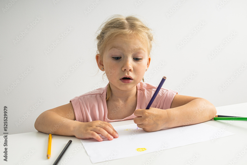 The child girl draws with colored pencils sitting at a white table on a white background The child girl draws with colored pencils sitting at a white table on a white background