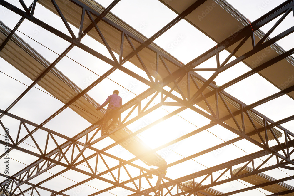 Two construction workers installing a new roof wear safety vests and safety harnesses to work on the steel roof structure.