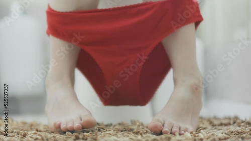 Woman sits on the toilet in a home bathroom, takes off her red panties and dangles her legs. Close-up of the legs. The concept of constipation, indigestion and diarrhea