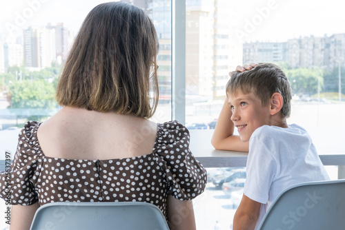 A cute happy little schoolboy boy is talking to his mother sitting at a table in a cafe against window. Mom talks to her son about school. The babysitter talks confidentially with the teenager. photo