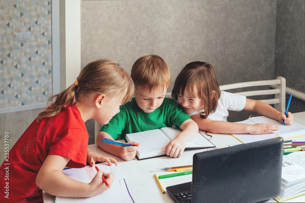 Children sit at a table with pens and notebooks and use a laptop