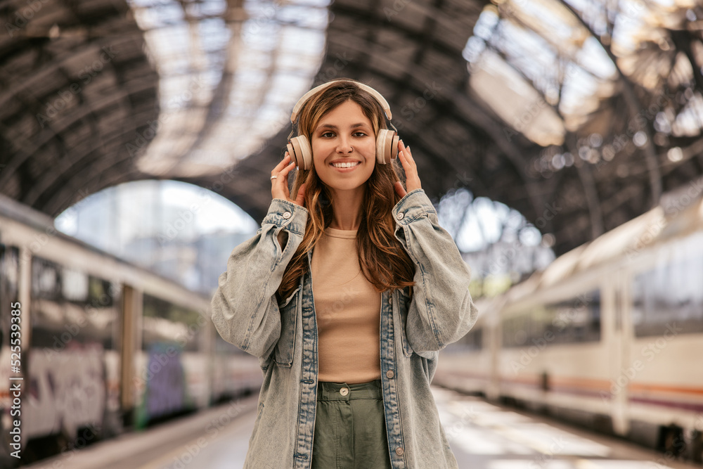 Beautiful young caucasian girl looks at camera, holds headphones on her head standing on station platform. Blonde wears casual clothes. Gadgets and music concept.