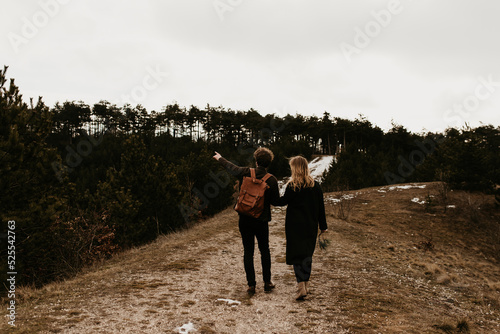 A couple is hiking in the mountains together. The man is pointing something in the distance. Late autumn, early winter tourist image with a couple in love. Trendy fashionable clothes, leather backpack