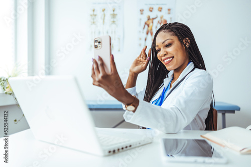 Headshot portrait of positive smiling African Amerian woman doctor in uniform sitting at desk videocalling at camera saying hi waving hand gesturing hello. Webcam view.  photo