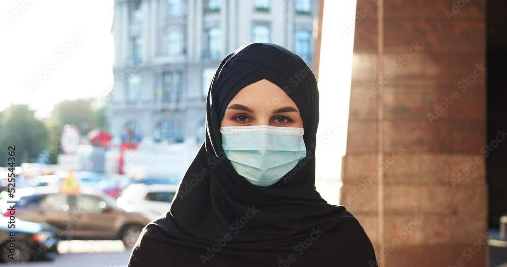 Young muslim Arab woman wearing hijab veiling face looking at camera outdoors during sunny day. Beautiful female Islam lady in medical mask covered with black headscarf. Religion, covid-19, virus. Young muslim Arab woman wearing hijab veiling face looking at camera outdoors during sunny day. Beautiful female Islam lady in medical mask covered with black headscarf. Religion, covid-19, virus.