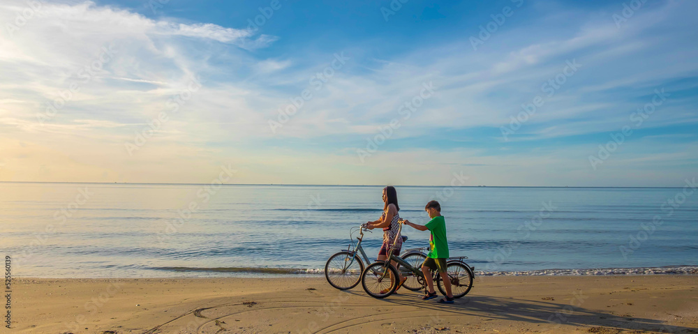 Fototapeta premium The lifestyle of a mother and son family on a bicycle at the sandy beach by the sea. The background image is a beautiful sunrise and sky. happy family concept