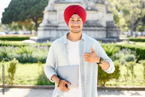 Positive smiling Indian man freelancer in turban showing thumbs up standing outdoors and holding laptop, satisfied hindu male student recommends studying on the distance from anywhere in the world photo