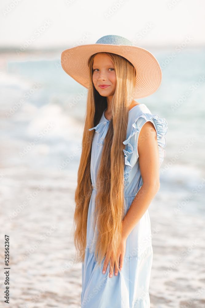 Beautiful smiling blonde teenager girl 12-14 year old with long hair wear straw hat and stylish elegant dress stand over sea coast outdoor. Summer season. Childhood.