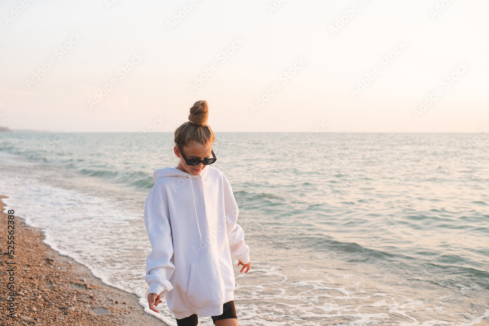 Stylish teenage girl 14-16 year old wear white cozy hoodie and hair bun with sun glasses walk over sea coast outdoor. Fashionable kid over water ocean. Summer vacation season.