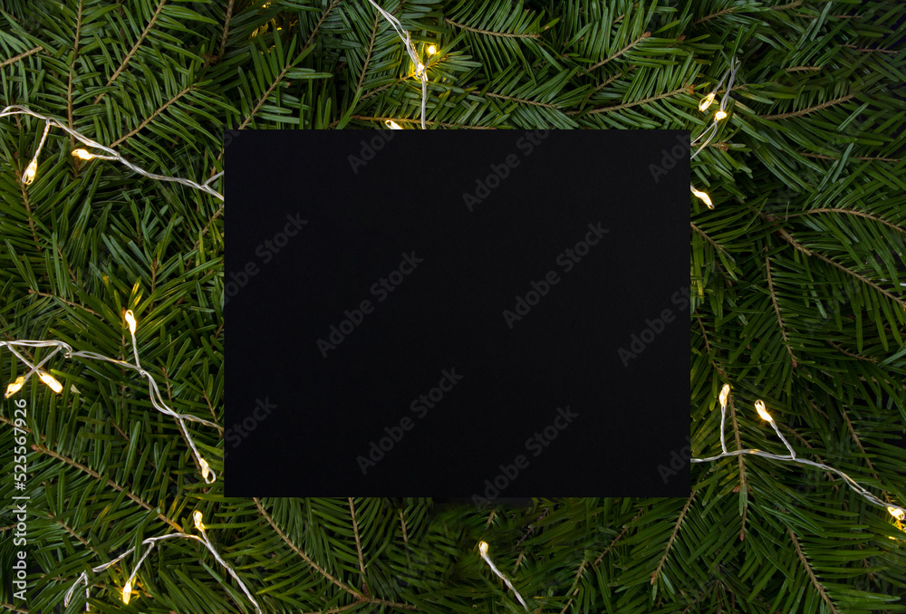 Christmas background. Close-up on fir branches with glowing garland and empty black card for text. Copy space.