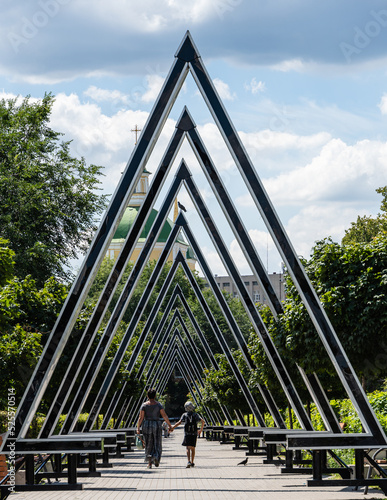 People walk down the original alley of architects with giant triangles. Green domes with golden Orthodox crosses of Church of Resurrection are in the distance. Voronezh, Russia - July 30, 2022 photo