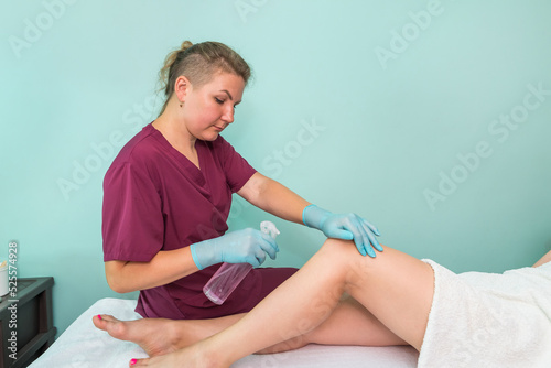 A beautician sprays a disinfectant on the feet of a young woman before the epilation procedure.
