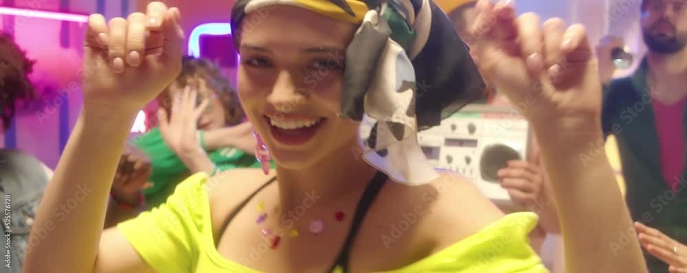 Young beautiful woman in bright outfit, headband and trendy jewelry dancing and smiling on camera at disco party with friends Young beautiful woman in bright outfit, headband and trendy jewelry dancing and smiling on camera at disco party with friends