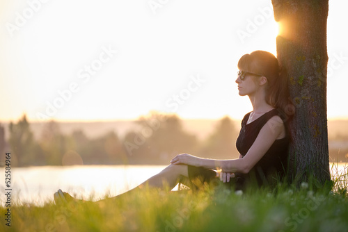 Photo Lonely young woman sitting alone on grass lawn on lake shore enjoying warm evening