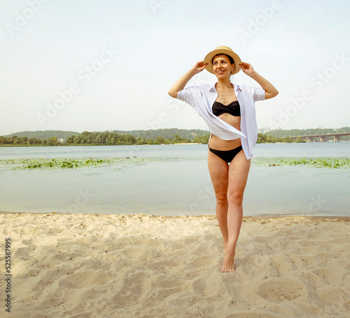 Wallpaper Mural A woman in a black swimsuit and a white shirt walks along the beach near the river in the city. Portrait of a woman in a hat near the water Torontodigital.ca