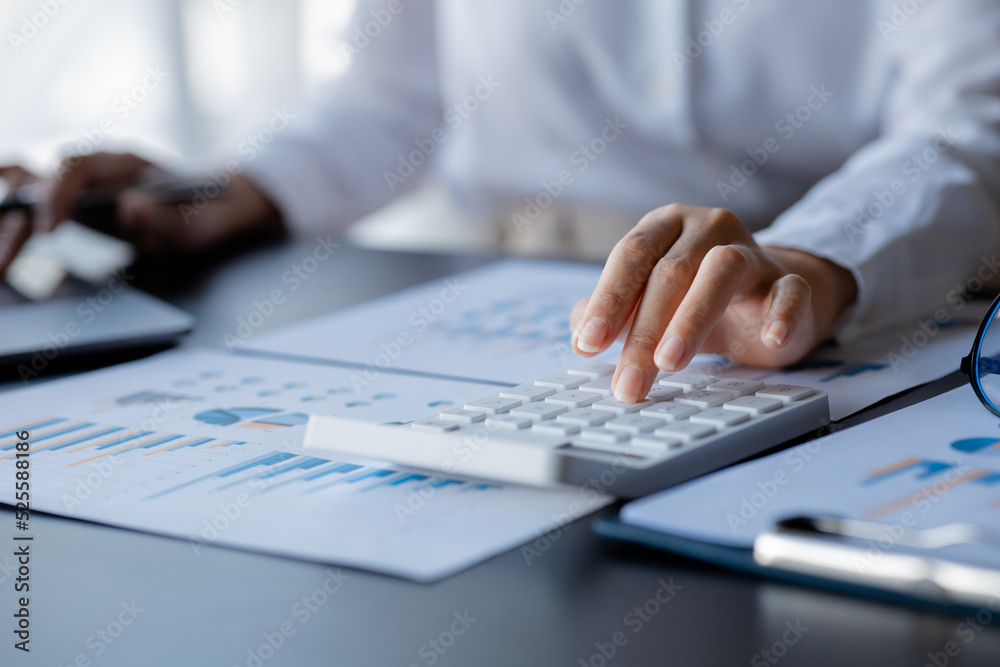 Businessman is using a calculator to calculate company financial figures from earnings papers, a businessman sitting in his office where the company financial chart is placed. Businessman is using a calculator to calculate company financial figures from earnings papers, a businessman sitting in his office where the company financial chart is placed.
