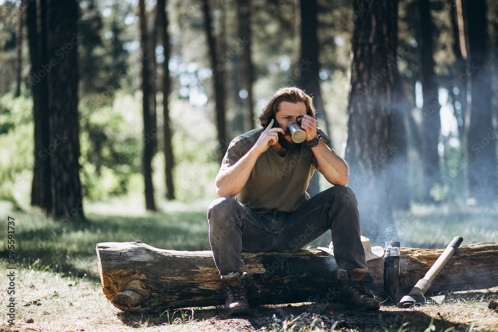 Fototapeta premium Man sits by fire in forest and drinks hot tea