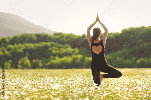 Fototapeta Naklejka Na Ścianę i Meble -  Young woman from behind doing yoga asana at nature landscape on backgroung. Active and sporty girl relaxing in chamomile field.