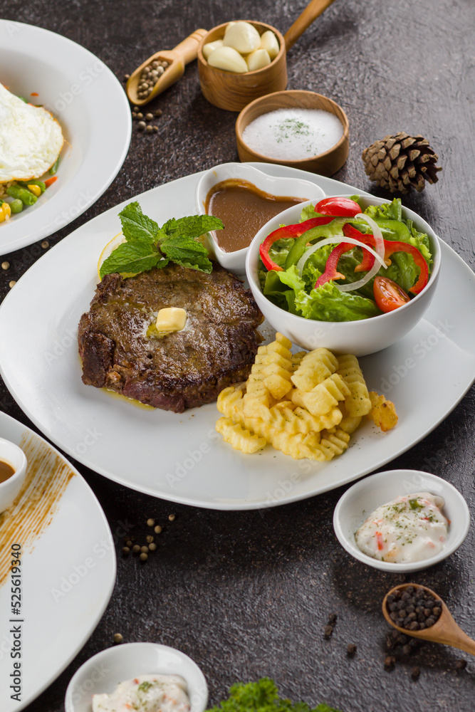 Grilled beef steaks with potato, vegie and steak sauce at white plate on texture rustic table