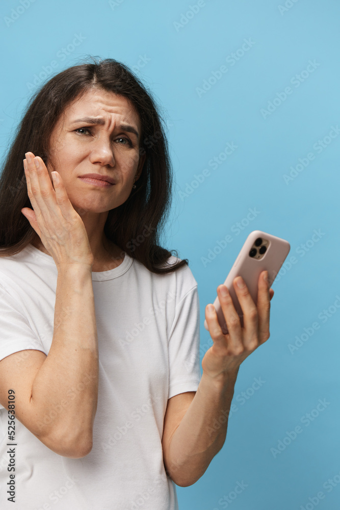 a very pensive woman stands on a blue background in a white T-shirt with a fashionable phone in her hands and looks at him in shock a very pensive woman stands on a blue background in a white T-shirt with a fashionable phone in her hands and looks at him in shock
