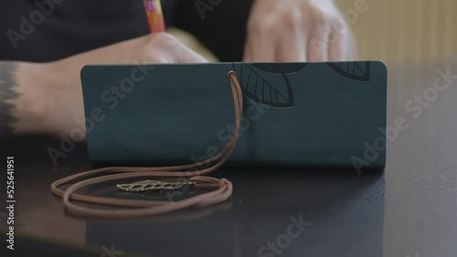 Hands With Tattooed Arm Writing in Creative Notebook with Pendant