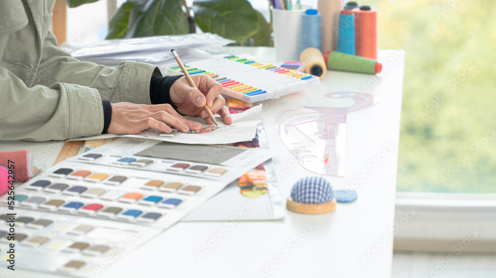 Selective focus on young hand of fashion designer, sketching or drawing creative clothes on paper on working desk at workplace with fabric color chart and tailor tools
