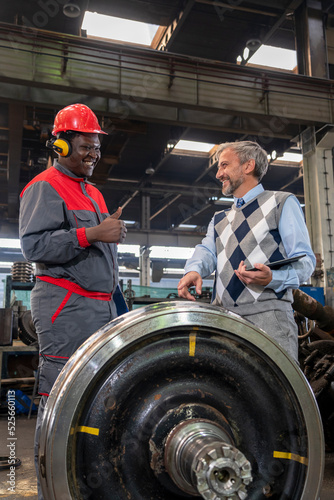 Smiling Black Worker In Protective Workwear And Smiling CEO Having A Relaxed Chat. Smiling Multiracial Industrial Co-Workers Standing In A Train Factory Next To Train Wheels. 