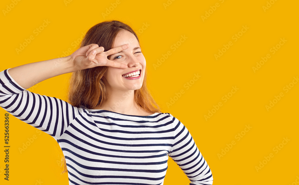 Studio shot of happy beautiful young woman. Cheerful Caucasian girl in striped top isolated on yellow background looks at camera, smiles, winks her eye and does peace, love and victory sign gesture