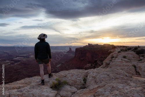 Adventurous Woman Hiking at a Desert Canyon with Red Rock Mountains. Cloudy Sunset Sky. Canyonlands National Park. Utah, United States. Adventure Travel
