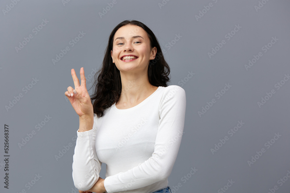 an excited woman stands on a gray background in a white T-shirt, joyfully shows a peace sign, putting her other hand on her stomach an excited woman stands on a gray background in a white T-shirt, joyfully shows a peace sign, putting her other hand on her stomach