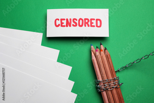 Card with word Censored, paper sheets, colorful pencils and chain on green background, flat lay photo