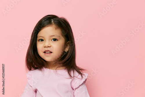 a cute little girl is standing on a pink background in a pink T-shirt with her hair loose and a pink strand, smiling admiringly showing her teeth and making faces turning her head in different