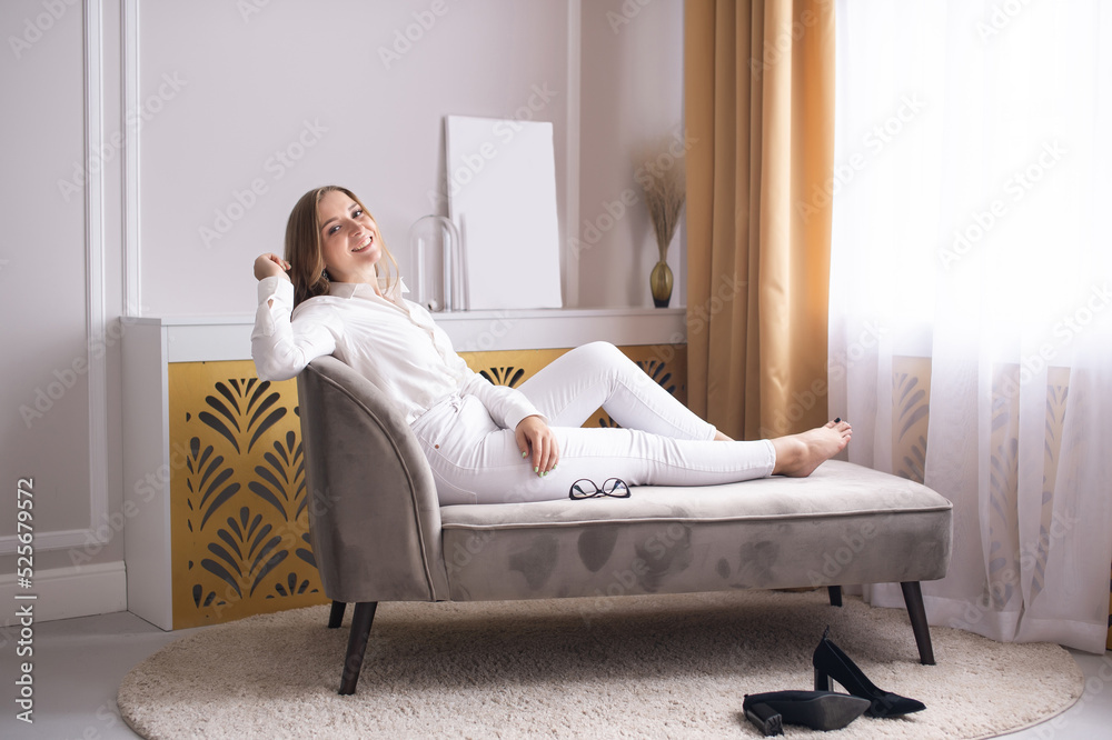 A cute girl in a white shirt is sitting on the sofa in the room and smiling