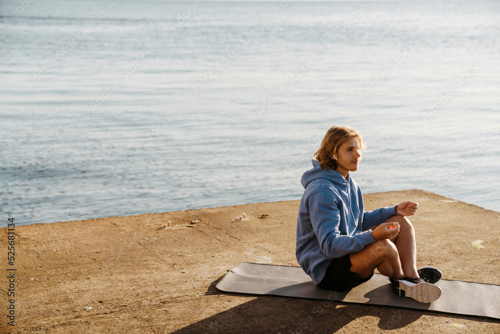 Young blonde man meditating on fitness mat while working out by sea Young blonde man meditating on fitness mat while working out by sea