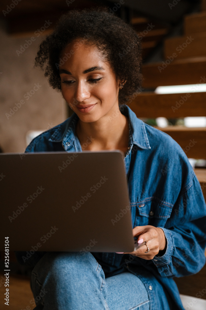 Young black woman wearing denim shirt smiling and using laptop at home