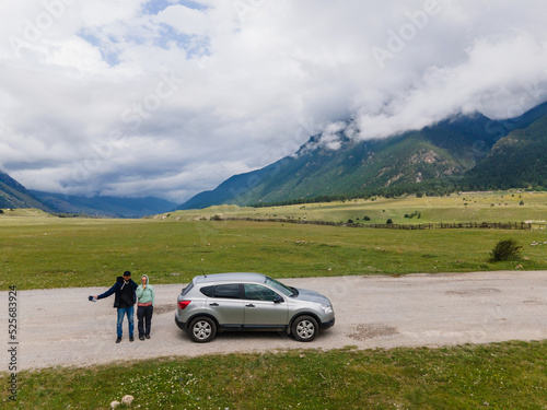 guy in a cap and jacket and a woman travelers are standing by the car among the Caucasus mountains in summer. go everywhere.