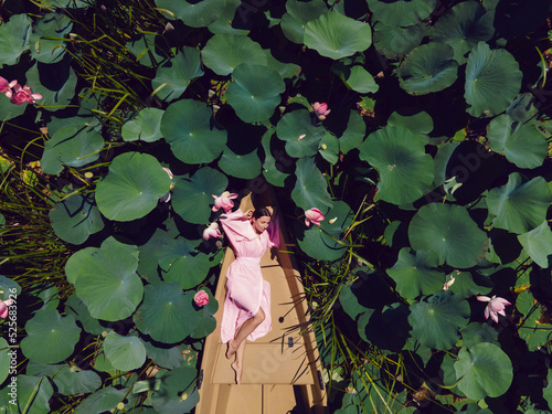 beautiful young fashionable woman in a pink dress lies on a wooden boat among blooming lotuses in summer. top view. go everywhere.