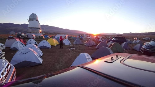 The time lapse of dawn in the camp near the observatory. The sun comes out from behind high hills. Red-yellow color. Lots of tents and cars. Guy launches the drone. Telescopes are covered with domes photo