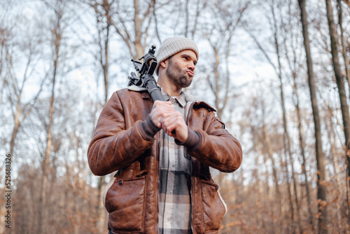 A portrait of a young white man posing in the woods dressed in autumn clothes: brown jacket, checkered canadian style shirt, off-white beanie, holding gimbal for videography as an ax