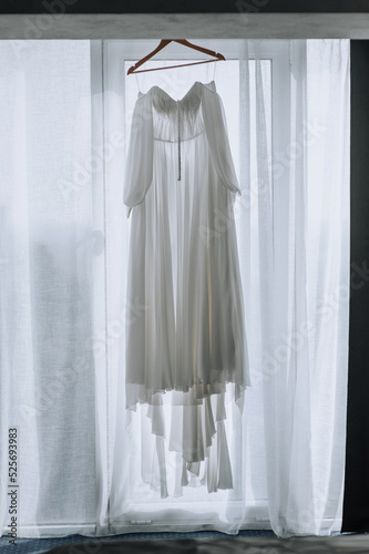 A beautiful long white bridesmaid dress hangs on a wooden hanger in front of the window. wedding photography.