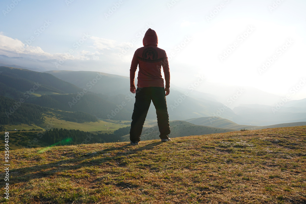 A guy on the edge of the hill admires the view. In the distance you can see high mountains and green hills covered with forest. Wide margins. Clouds are visible and sun is shining brightly. Kazakhstan