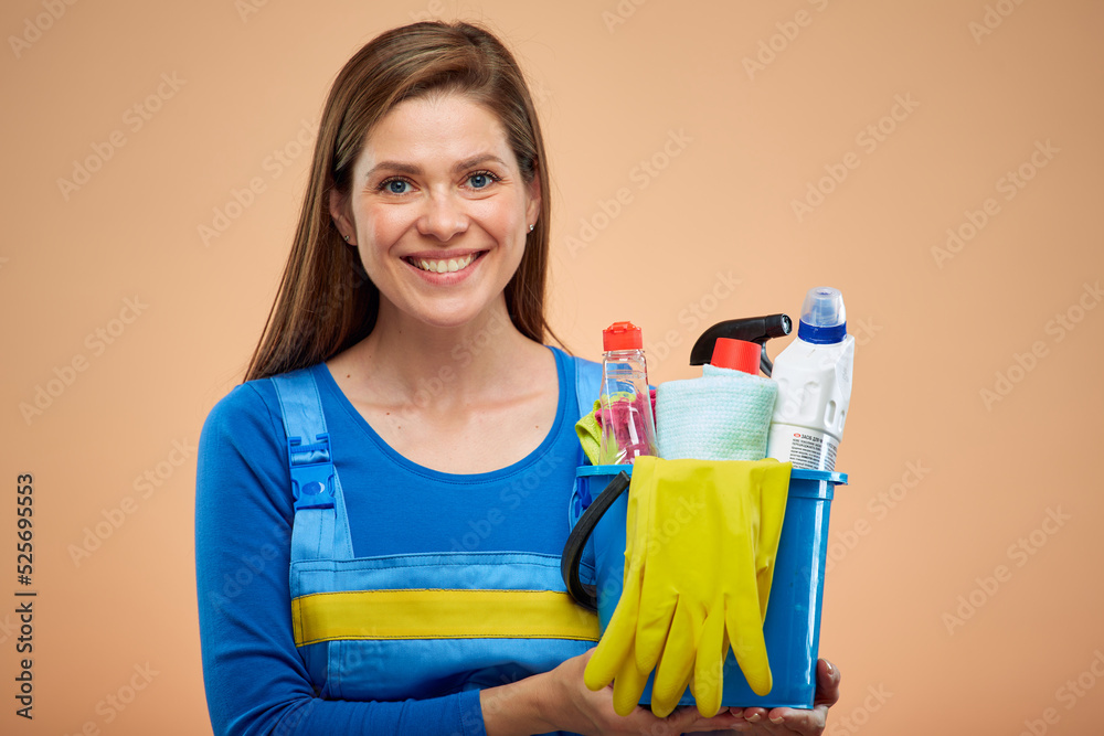 Woman cleaner in overalls holding cleaning products in bucket. isolated female housekeeper portrait on brown beige background. Woman cleaner in overalls holding cleaning products in bucket. isolated female housekeeper portrait on brown beige background.