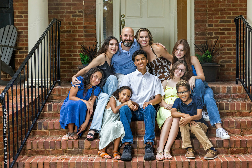 A large and blended and mixed happy family sitting on the front steps of their brick home in the suburbs - obrazy, fototapety, plakaty 