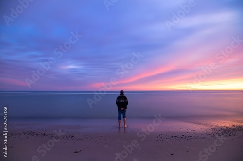 Obraz na plátně Back shot of an adult man stands at the beach looking at the sea and dusk sky